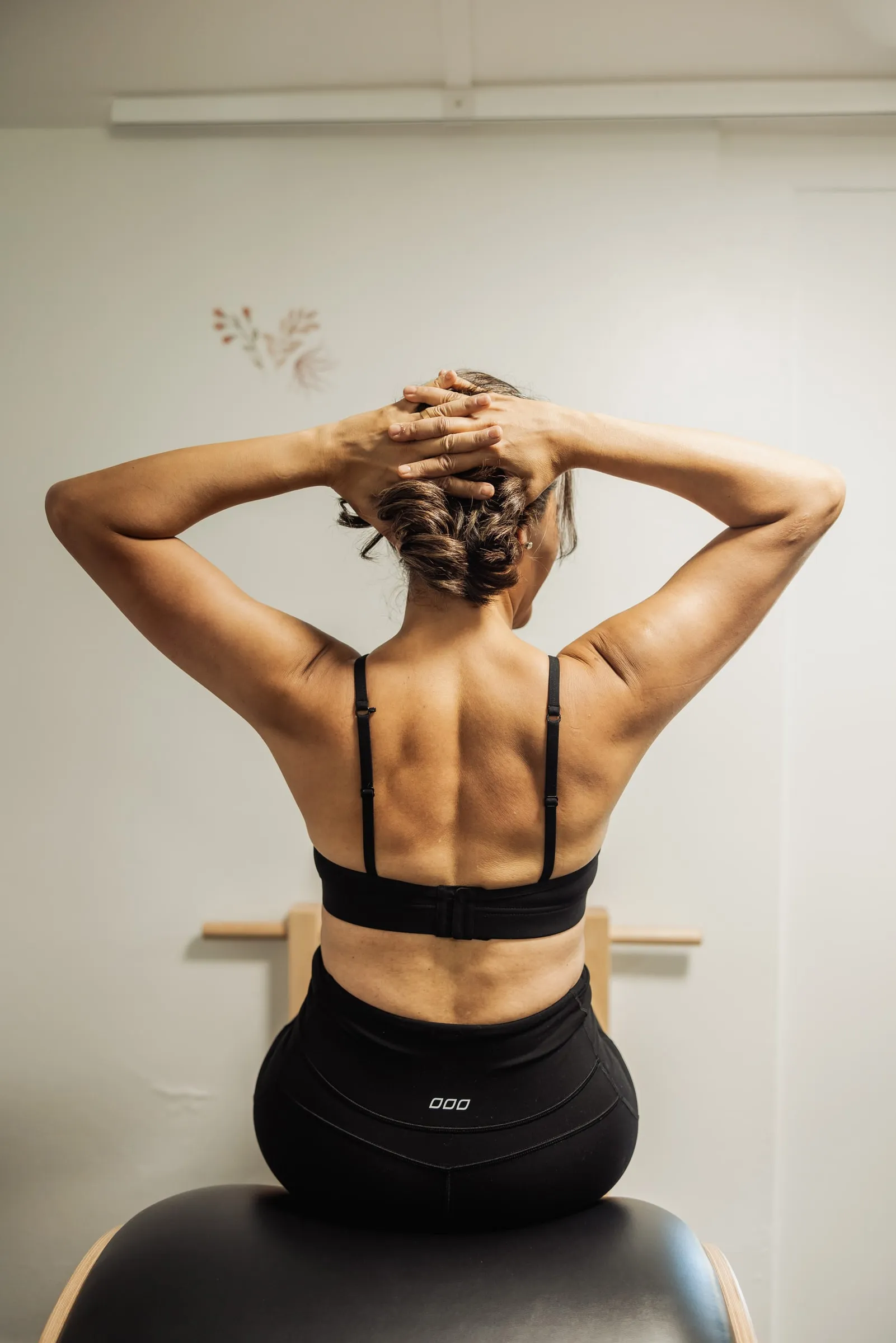 Meher seated on a Pilates barrel with her hands behind her head, working on posture and alignment